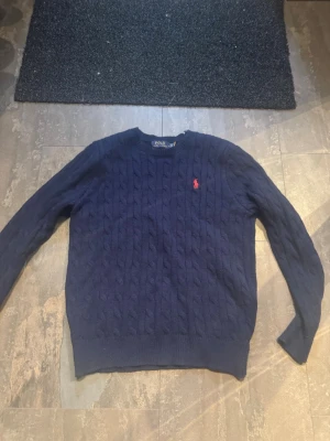 Polo Ralph lauren stickad tröja  - Kabelstickad mörkblå tröja från Polo Ralph Lauren med rund halsringning och den klassiska röda logotypen broderad på bröstet. Tröjan har långa ärmar och ribbade muddar vid ärmslut och nederkant. Perfekt för dig som gillar stilrena och tidlösa plagg.