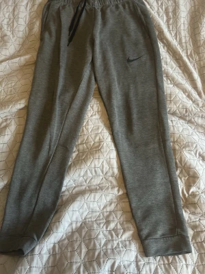 Gråa mjukisbyxor från Nike - Snygga gråa mjukisbyxor från Nike med svart logga på vänster ben. Byxorna har dragsko i midjan och muddar vid bensluten för en skön passform. Tillverkade i mjukt material som passar perfekt för chill eller träning.