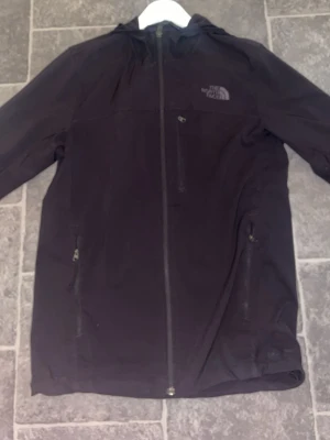 Svart vindjacka från The North Face - Snygg svart vindjacka från The North Face med huva och hel dragkedja framtill. Jackan har tre fickor med dragkedja och diskret logga på bröstet och ryggen. Tillverkad i lätt och tåligt material, perfekt för blåsiga dagar.