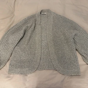 Grå stickad kofta  - Mysig grå stickad kofta från Vero Moda med öppen front och bred ribbad kant. Koftan har en avslappnad passform och långa ärmar med strukturstickat mönster. Perfekt att slänga över en t-shirt eller topp för en chill look.