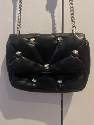 Svart axelväska med nitar och kedja - Snygg svart axelväska med quiltad design och silverfärgade nitar. Väskan har en axelrem i kedja och läder, samt ett lock med dekorativa detaljer. Perfekt för dig som gillar en edgy och trendig stil.