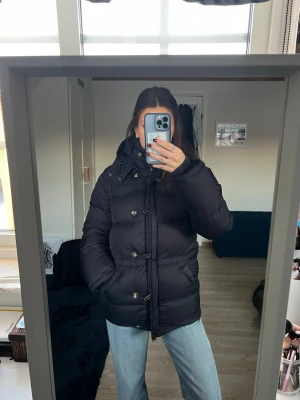 Mörkblå dunjacka med huva - Snygg mörkblå dunjacka med huva och knappar framtill från Bommerang i storlek xs. Jackan är lång, har en pufferstil och stora fickor på sidorna. Perfekt för kalla dagar och har en klassisk look som passar till det mesta. Hade ett litet hål vid ärmen som är ihop sytt. Inga andra defekter bortsett från de. Nypris: 7000kr.🤗🤗