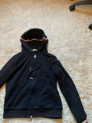 Mörkblå hoodie med dragkedja - Snygg mörkblå hoodie från Moncler med dragkedja framtill och fickor. Huvan har coola ränder i vitt, rött och gult samt dragsko med metalländar. Perfekt för en avslappnad streetwear-look. Tillverkad i mjukt bomullsmaterial.Xs