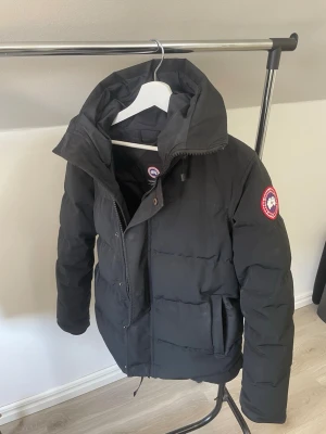 Svart vinter jacka från Canada Goose - Säljer en svart dunjacka från Canada Goose med hög krage och klassisk logotyp på ärmen. Jackan har dragkedja och tryckknappar framtill samt två stora fickor. Perfekt för kalla vinterdagar och riktigt snygg streetstil.