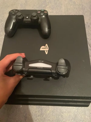 PlayStation 4 Pro med två kontroller - Säljer en svart PlayStation 4 Pro från Sony med två matchande DualShock 4-kontroller. Konsolen har den klassiska fyrkantiga designen med PlayStation-loggan i silver på ovansidan. Perfekt för gaming och streaming, koppla in och kör igång direkt!