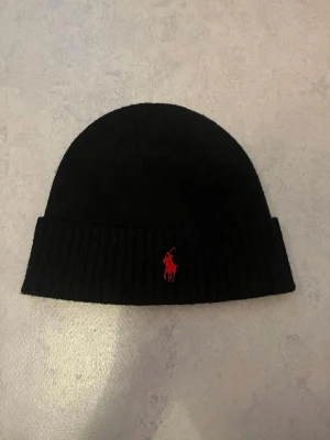 Svart mössa från Polo Ralph Lauren - Snygg svart mössa från Polo Ralph Lauren i mjuk merinoull. Klassisk ribbad kant och röd broderad logga framtill. Perfekt för kalla dagar och enkel att matcha med olika outfits.