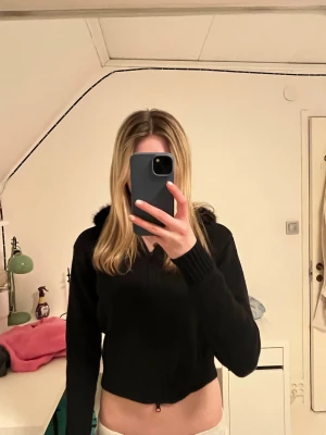 Svart hoodie med fuskpäls på huvan - Snygg svart hoodie med dragkedja och stor huva kantad med fuskpäls. Croppad modell med ribbade muddar vid ärmslut och midja. Perfekt för dig som vill ha en trendig och mysig look.