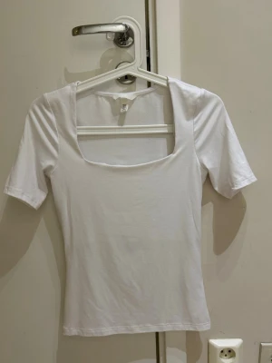  Ny Vit topp med fyrkantig ringning - Säljer en enkel och stilren vit topp med fyrkantig ringning och korta ärmar. Toppen är gjord i mjuk bomull och har en tight passform som sitter snyggt på kroppen. Perfekt att matcha med jeans eller kjol för en clean look.