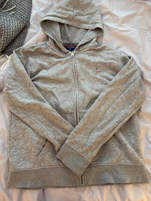 Grå zip hoodie från Gant - gråmelerad hoodie från Gant med dragkedja framtill och broderad logga på bröstet. Tröjan har huva och två fickor fram. Tillverkad i mjuk bomullsblandning