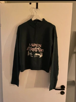 Grön stickad tröja Boohoo Aspen - Mörkgrön stickad tröja från Boohoo med halv dragkedja och hög krage. Framtill står det 'Aspen Apres Ski' i vitt och rosa med vintermotiv. Perfekt för kalla dagar och har en avslappnad passform. Finns på fler sidor 