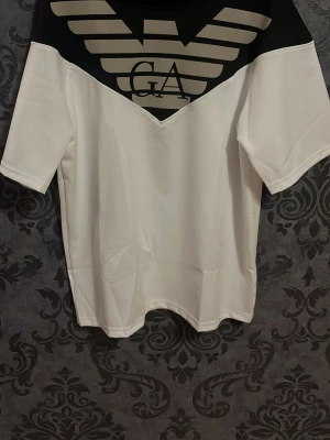 Vit och svart t-shirt från Armani - Snygg t-shirt från Armani med vit bas och svart parti upptill. Stort ikoniskt logotryck i beige och svart över bröstet. Klassisk passform med korta ärmar och rund hals. Perfekt för dig som vill sticka ut med en stilren look.