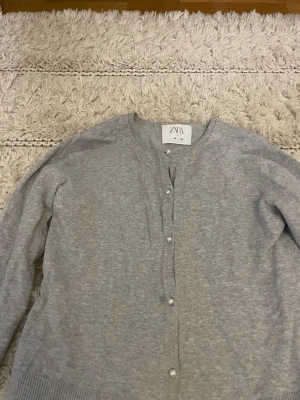 Grå kofta från Zara med pärlknappar - Snygg grå kofta från Zara med rund halsringning och dekorativa pärlknappar framtill. Koftan har ribbade muddar vid ärmslut och nederkant samt en klassisk, enkel design som passar till det mesta.