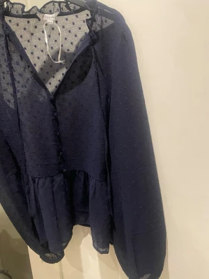 Mörkblå prickig blus Happy Holly - Säljer en mörkblå blus från Happy Holly med transparenta partier och små prickar i tyget. Blusen har v-ringning, knappar framtill och volangdetalj nedtill. Långa ärmar med resår vid ärmslut ger en luftig och trendig look. Helt ny aldrig använd dock utan pris lapp 