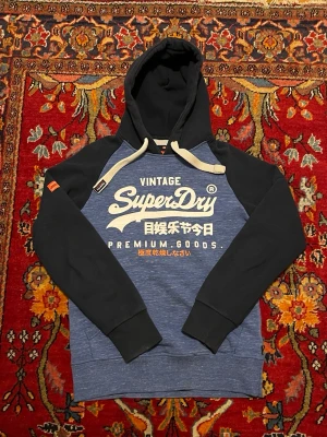 Blå Superdry hoodie med tryck - Snygg hoodie från Superdry i blått med mörkblå ärmar och huva. Stor vit logga och japansk text på bröstet. Dragsko i huvan och ribbade muddar. Perfekt för en avslappnad streetwear-look.
