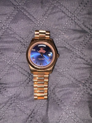 Rosé guld  klocka med blå urtavla - Snygg rosé guld klocka från R med blå urtavla och romerska siffror. Klockan har en klassisk länkarmband i metall och dag- och datumvisning. Perfekt accessoar för att lyfta din stil. Rosé guld färg 