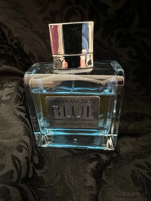 San Francisco Blue Eau de Toilette 100ml - San Francisco Blue for Men är en eau de toilette i en stilren, rektangulär glasflaska med blått innehåll och silverfärgad etikett. Flaskan har en blank, silverfärgad kork och rymmer 100 ml. Doften är maskulin och modern, perfekt för dig som gillar fräscha och eleganta parfymer.