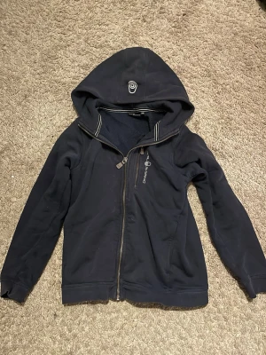 Sail racing hoddie - Mörkblå hoodie med dragkedja från Sail racing. Utrustad med huva som har ett vitt tryck framtill och vita detaljer längs dragkedjan. Insidan är mjuk och fleecefodrad för extra komfort. Perfekt för en avslappnad och sportig stil.