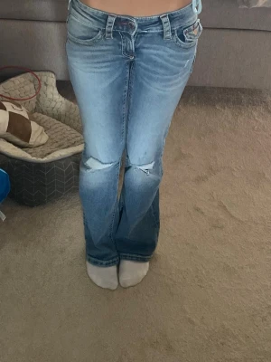 Bootcut jeans med slitningar - Säljer ett par ljusblå bootcut jeans med slitningar vid knäna och snygga detaljer på bakfickorna. Jeansen har låg midja och klassisk femficksdesign. Perfekta för en avslappnad och trendig look.