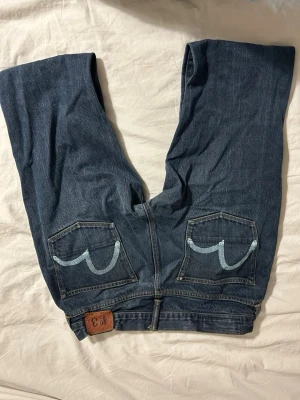 Evisu jeans med broderade bakfickor - Säljer ett par klassiska blå jeans från Evisu med deras ikoniska målad båge på bakfickorna. Jeansen är raka i modellen och har detaljer i läder vid linningen. Tillverkade i slitstark denim av hög kvalitet. Perfekta för dig som gillar streetwear och japansk design.