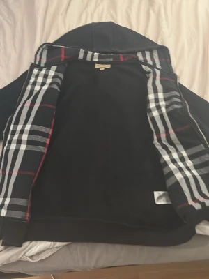 Svart zip-hoodie från Burberry - Snygg svart hoodie med dragkedja från Burberry. Klassisk huva och två fickor framtill. Insidan har det ikoniska Burberry-rutiga fodret i svart, vitt och rött. Perfekt för dig som vill ha en stilren och exklusiv look. Storlek M men passar S