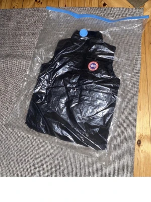 Svart dunväst från Canada Goose - Snygg svart dunväst från Canada Goose med klassisk logotyp på bröstet. Västen har hög krage och är tillverkad i ett glansigt material som ger en stilren look. Perfekt att bära över en hoodie eller tröja för extra värme.