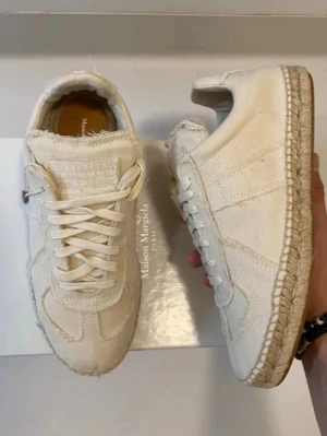 Maison Margiela vita sneakers i canvas - Unika vita sneakers från Maison Margiela i canvas med detaljer i mocka. Skorna har klassisk snörning, rund tå och en sula med espadrille-inspirerad flätning. Perfekt för dig som gillar stilrena och trendiga sneakers med en twist. Står 38 men sitter som 40