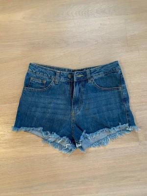 Blå korta jeansshorts med fransig kant från Nelly - Säljer ett par supersnygga klassiska korta blå jeansshorts från Nelly Jeans. Shortsen har en fransig kant nedtill och ger en avslappnad look. Perfekta för varma dagar och enkla att matcha med olika toppar. Säljer då jag har ett par andra jag använder mer