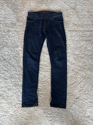 Mörkblå jeans från Nudie Jeans Grim Tim - Snygga mörkblå jeans från Nudie Jeans i modellen grim tim med klassisk femficksdesign och raka ben. Jeansen har kontrastsömmar och är tillverkade i denim med en stilren look. Perfekta för dig som gillar en tidlös och enkel stil.
