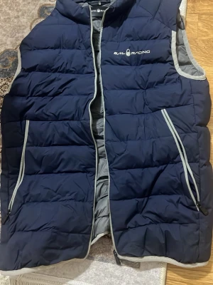 Marinblå dunväst från Sail Racing - Snygg marinblå dunväst från Sail Racing med grå detaljer och dragkedja framtill. Västen har två snedställda fickor med dragkedja och logga på bröstet. Insidan är grå och quiltad för extra värme. Perfekt lager på lager-plagg för kyliga dagar.