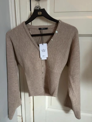 Beige kofta från Gina Tricot - Trendig beige kofta från Gina Tricot med ribbad midja och v-ringning. Koftan har långa ärmar, knappar framtill och en croppad passform som ger en snygg siluett. Perfekt att styla med höga jeans eller kjol.