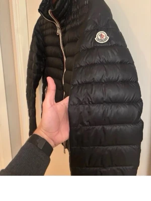 Svart dunjacka från Moncler - Snygg svart dunjacka från Moncler med quiltad design och hög krage. Jackan har två framfickor med dragkedja och Moncler-logga på ärmen. Dragkedjan framtill har en vit och röd detalj som ger en sportig touch. Perfekt för kalla vinterdagar. Kvitto finns 