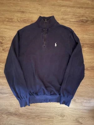 Mörkblå halvzip från Polo Golf - Mörkblå stickad tröja från Polo Golf Ralph Lauren med halvzip och vit broderad logga på bröstet. Tröjan har ribbade muddar, hög krage med vit insida och är perfekt för lager på lager. Klassisk och stilren design.
