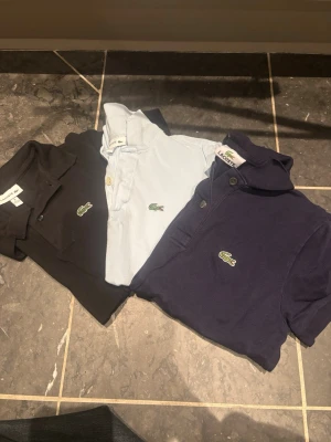 3-pack pikétröjor från Lacoste - Säljer tre klassiska pikétröjor från Lacoste i svart, ljusblå och marinblå. Alla har den ikoniska krokodilloggan på bröstet och krage med knappar. Perfekta för en clean och sportig stil. Materialet är mjuk bomull som andas.