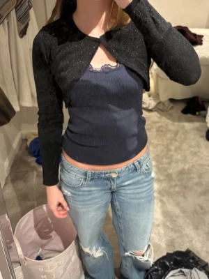 Svart glittrig bolero med lång ärm - Kort svart bolero med glittrigt tyg och lång ärm. Stängs framtill med en hake. Perfekt att slänga över ett vitt linne eller klänning för en snygg layering-effekt.