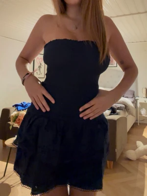 Marin blå axelbandslös kort klänning - Säljer en marin blå axelbandslös kort klänning med smock upptill och volangig kjol. Klänningen har en feminin siluett och är perfekt för dig som vill ha en enkel men snygg look. Materialet känns lätt och passar bra till varma dagar.