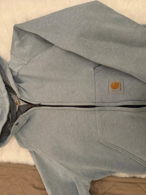 Ljusgrå hoodie från Carhartt - Snygg ljusgrå hoodie från Carhartt med dragkedja framtill och klassisk huva. Hoodien har en stor ficka på magen och Carhartt-logga på bröstet. Tillverkad i mjukt bomullsmaterial som känns skönt mot huden. Perfekt för en avslappnad streetwear-look.