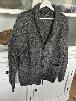 Gråmelerad kofta med knappar - Mysig gråmelerad kofta med grovstickad känsla och stora bruna knappar framtill. Koftan har två framfickor och bred krage som ger en avslappnad look. Perfekt för lager på lager och chill dagar.