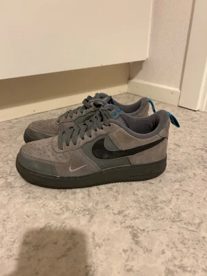Grå Nike Air Force 1 sneakers - Snygga grå Nike Air Force 1 sneakers i mocka och läder med svart swoosh och mörkgrå sula. Skorna har blå detaljer på plösen och hälen samt klassisk låg siluett. Perfekta för dig som gillar stilrena och tidlösa sneakers.