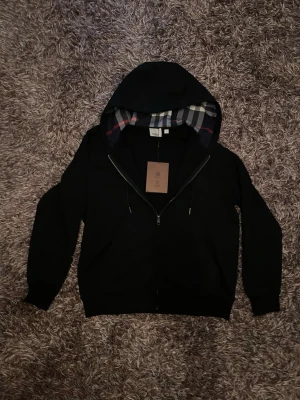 Svart hoodie från Burberry - Säljer en svart hoodie från Burberry med dragkedja och klassiskt rutigt foder i huvan. Hoodien har långa ärmar, ribbade muddar och en liten Burberry-logga på bröstet. Perfekt för dig som gillar stilrena och exklusiva plagg. Förpackningen följer med också!