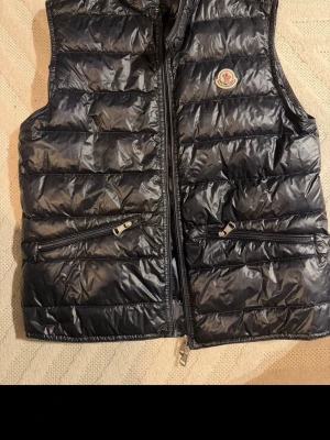 Svart dunväst från Moncler - Snygg svart dunväst från Moncler med glansig finish och klassisk logga på bröstet. Västen har två dragkedjefickor framtill och quiltad design. Perfekt för lager-på-lager och håller dig varm med stil.