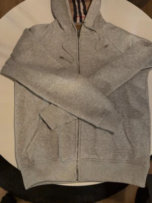 Grå zip hoodie från Burberry - Snygg grå zip hoodie från Burberry med klassiskt broderat riddarlogo på bröstet och den ikoniska rutiga insidan av huvan. Hoodien har dragsko i huvan, fickor fram och ribbade muddar. Perfekt för en chill och stilren look.