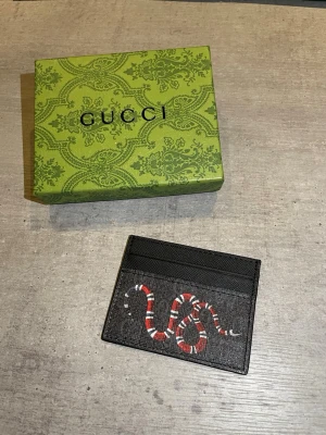  svart korthållare med orm - Snygg svart korthållare med ikoniskt ormmotiv i rött, vitt och svart. Tillverkad i exklusivt läder med plats för flera kort. Kommer med original grön box med mönster. Perfekt accessoar för dig som gillar lyxiga detaljer.