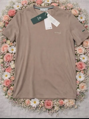 Beige t-shirt från Lacoste - Snygg beige t-shirt från Lacoste med diskret logga på bröstet och det klassiska krokodilmärket på ärmen. T-shirten har rund halsringning och korta ärmar. Tillverkad i mjuk bomull för en skön känsla och clean look.