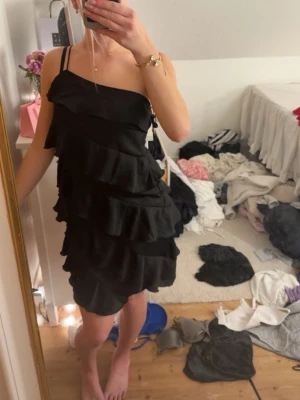 Svart volangklänning från Bubbleroom - Supersnygg svart kort klänning från Bubbleroom med flera lager volanger och tunna axelband. Klänningen har en asymmetrisk one shoulder-design och är perfekt för dig som vill sticka ut. Materialet känns lätt och följsamt.