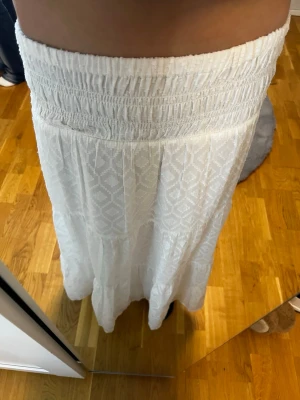 Vit långkjol med mönster - Säljer en vit långkjol med diskret geometriskt mönster och bred, smockad midja. Kjolen är luftig och har en lätt volang nertill, perfekt för varma dagar. Superfin att styla med en enkel topp eller stickad tröja.