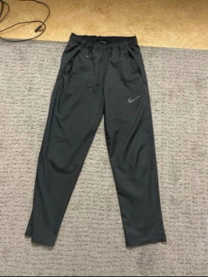 Svarta mjukisbyxor från Nike - Svarta mjukisbyxor från Nike med dragsko i midjan och diskret Nike-logga på ena benet. Byxorna har raka ben och är tillverkade i ett mjukt och lätt material som passar perfekt för chill eller träning.