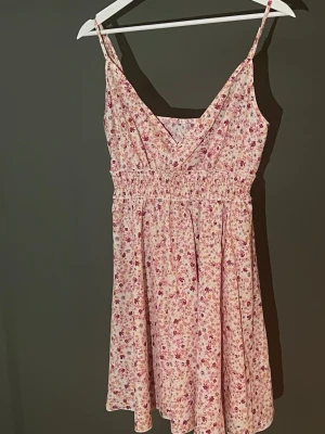 Blommig kort sommarklänning - Söt kort klänning med smala axelband och v-ringning. Klänningen är vit med små rosa och röda blommor, har resår i midjan och ett luftigt fall. Perfekt för varma dagar och ger en romantisk vibe.