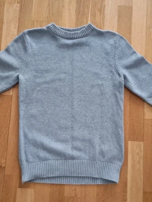 Ljusgrå stickad tröja - En stilren ljusgrå stickad tröja med rund halsringning och ribbade muddar vid ärmslut och nederkant. Tröjan har en klassisk passform och är perfekt för lager-på-lager. 