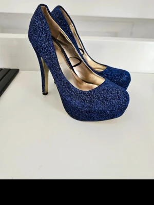 Blå glittriga pumps från Centro - Säljer ett par blå glittriga pumps från Centro med hög platå och smal klack. Skorna har rundad tå och insida i guldigt material. Perfekta för dig som vill sticka ut med extra höjd och glitter. Se bilder för Skavanker. Storlek 38. Passar inte breda fötter!