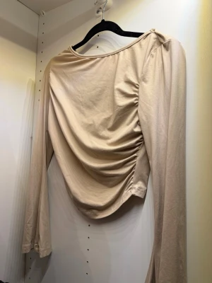 Beige croppad topp med rynk - Trendig beige croppad topp med långa ärmar och snygg rynkdetalj på sidan. Toppen har en bred, rund halsringning och är tillverkad i mjukt bomullsmaterial. Perfekt för dig som gillar stilrena och moderna plagg.